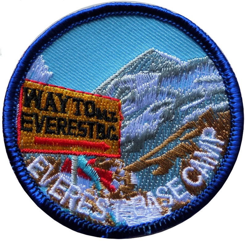 May include: A blue and white embroidered patch featuring a mountain scene with the text "WAY TO EVEREST BASE CAMP" in white and gold lettering. The text "WAY TO EVEREST" is on a yellow sign with a red border.