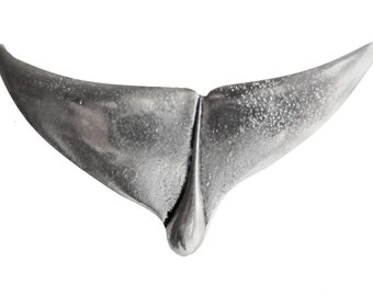 May include: A silver-colored whale tail pendant, isolated on a white background. The pendant has a textured surface and a split design, with a rounded base. The tail's flukes curve upwards.