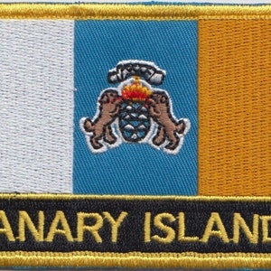 May include: A yellow, white and blue embroidered patch with the text "CANARY ISLANDS" in black lettering. The patch features a coat of arms with two dogs and a pine cone.