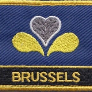 May include: A blue embroidered patch featuring a yellow and grey heart-shaped symbol and the word "BRUSSELS" in yellow lettering on a black background.
