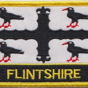 Op de afbeelding: Een witte patch met een zwart kruis en vier zwarte vogels met rode snavels en poten. De tekst "FLINTSHIRE" staat in gele letters op een zwarte achtergrond.