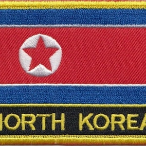 Könnte beinhalten: Ein Nordkorea-Flaggen-Patch mit rotem Hintergrund, einem blauen Streifen oben und unten und einem weißen Stern in einem roten Kreis in der Mitte. Der Patch hat einen schwarzen Rand mit der Aufschrift "NORTH KOREA" in Gelb.