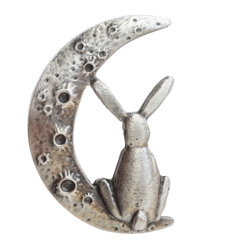 May include: A silver-toned metal charm featuring a rabbit sitting on a crescent moon. The moon has a textured surface with small circles representing craters.