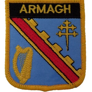 May include: Embroidered patch featuring the word "ARMAGH" in gold on a black banner. The patch has a light blue background with a red diagonal stripe and gold accents, including a harp and a cross symbol. The patch is bordered with gold stitching.