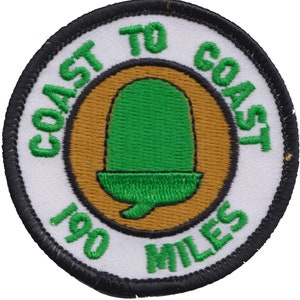 Può includere: Un cerotto rotondo bianco con un bordo nero. Al centro del cerotto c'è un cerchio marrone con una ghianda verde al suo interno. Il testo "COAST TO COAST 100 MILES" è scritto in verde attorno al cerchio marrone.