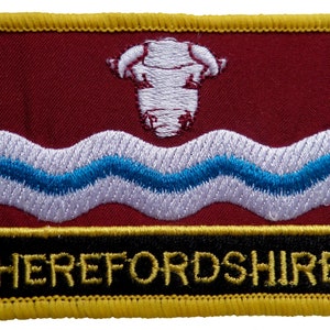 May include: An embroidered patch featuring a white bull's head on a red background with white and blue wavy lines. The text "HEREFORDSHIRE" is in yellow lettering on a black background.