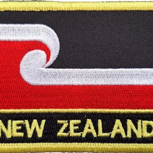 Könnte beinhalten: Ein gestickter Aufnäher in Schwarz, Rot und Weiß mit einem stilisierten Wellendesign und dem Text "NEW ZEALAND" in Gelb.