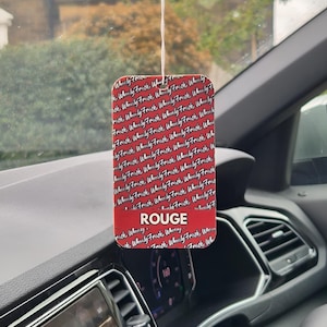 May include: A red rectangular car air freshener with the word "ROUGE" in white. The air freshener has a repeating pattern of the words "Wheely Fresh" in white. It hangs from a string inside a car.