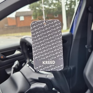 May include: Rectangular car air freshener with the word "KREED" in white text. The air freshener is black with the words "Wheely Fresh" in a repeating pattern. The air freshener is hanging in a car.