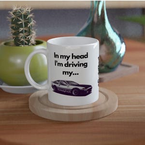 May include: White ceramic mug featuring the text "In my head I'm driving my..." and a black and white car graphic. The mug rests on a wooden coaster. A green potted cactus and a decorative vase are in the background.