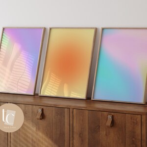 May include: Three framed abstract art prints with a gradient of pastel colors. The prints are in wooden frames and are displayed on a wooden cabinet.