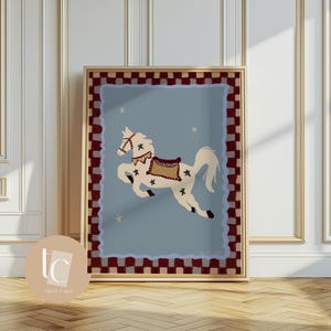 May include: A framed art print featuring a whimsical illustration of a white carousel horse with a brown saddle and star accents, set against a light blue background. The artwork is framed with a gold border and a red and white checkered pattern.