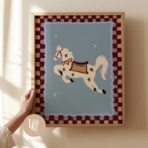 May include: A framed art print featuring a whimsical illustration of a white horse with a saddle and star accents against a light blue background. The artwork is framed with a gold-colored border and a red and white checkered pattern.