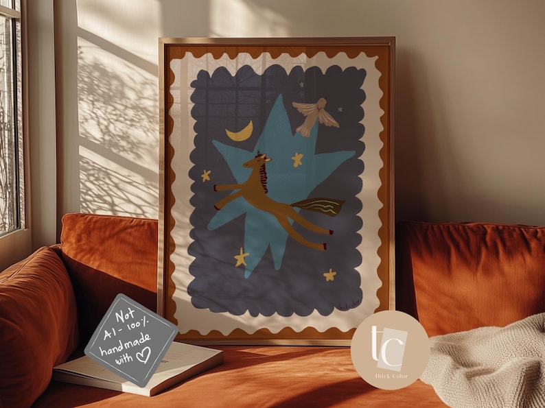 May include: A framed art print featuring a whimsical illustration of a brown unicorn soaring through a blue star-shaped design, with a crescent moon and stars. The artwork has a beige border and is displayed on a rust-colored couch.