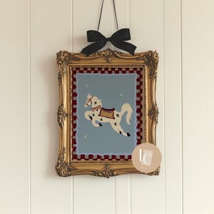 May include: A framed artwork featuring a white horse with star accents on a light blue background. The horse is adorned with a saddle and bridle. The gold frame is ornate, and a black bow suspends the artwork.