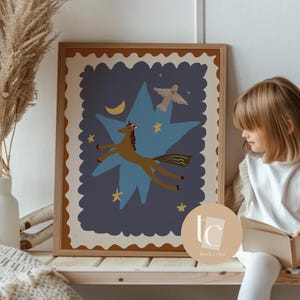 May include: A framed art print featuring a whimsical illustration of a brown unicorn soaring through a blue star-shaped design, with a crescent moon and stars. The artwork has a beige border and is displayed on a wooden shelf.