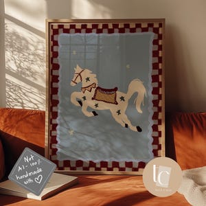 May include: A framed art print featuring a whimsical illustration of a carousel horse. The horse is cream-colored with a decorative saddle and star accents, set against a light blue background. The frame has a red and white checkered border.
