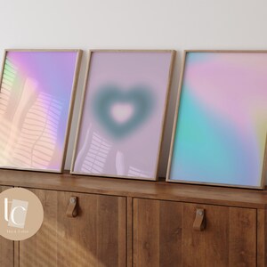 May include: Three framed abstract art prints with a gradient of pink, blue, and green colors. The middle print features a heart shape in the center.