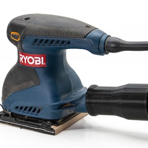Op de afbeelding: Een blauwe en zwarte Ryobi elektrische schuurmachine met een stofafzuigbuis. De schuurmachine heeft een rechthoekig schuurplateau en een trekker.