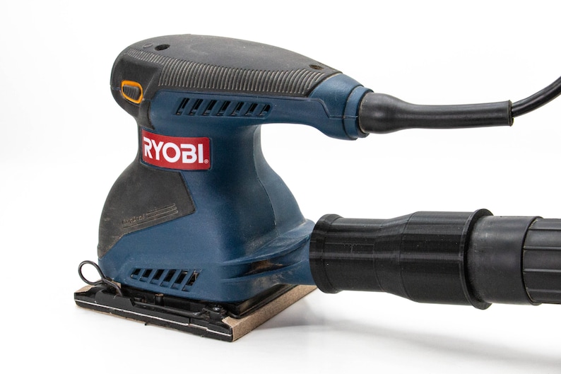 Op de afbeelding: Een blauwe en zwarte Ryobi elektrische schuurmachine met een stofafzuigbuis. De schuurmachine heeft een rechthoekig schuurplateau en een trekker.