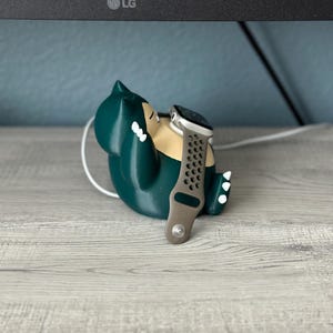 Puede incluir: Un personaje de Pokemon impreso en 3D de color verde, que se asemeja a un Snorlax, sostiene un Apple Watch plateado con una banda deportiva negra y gris.