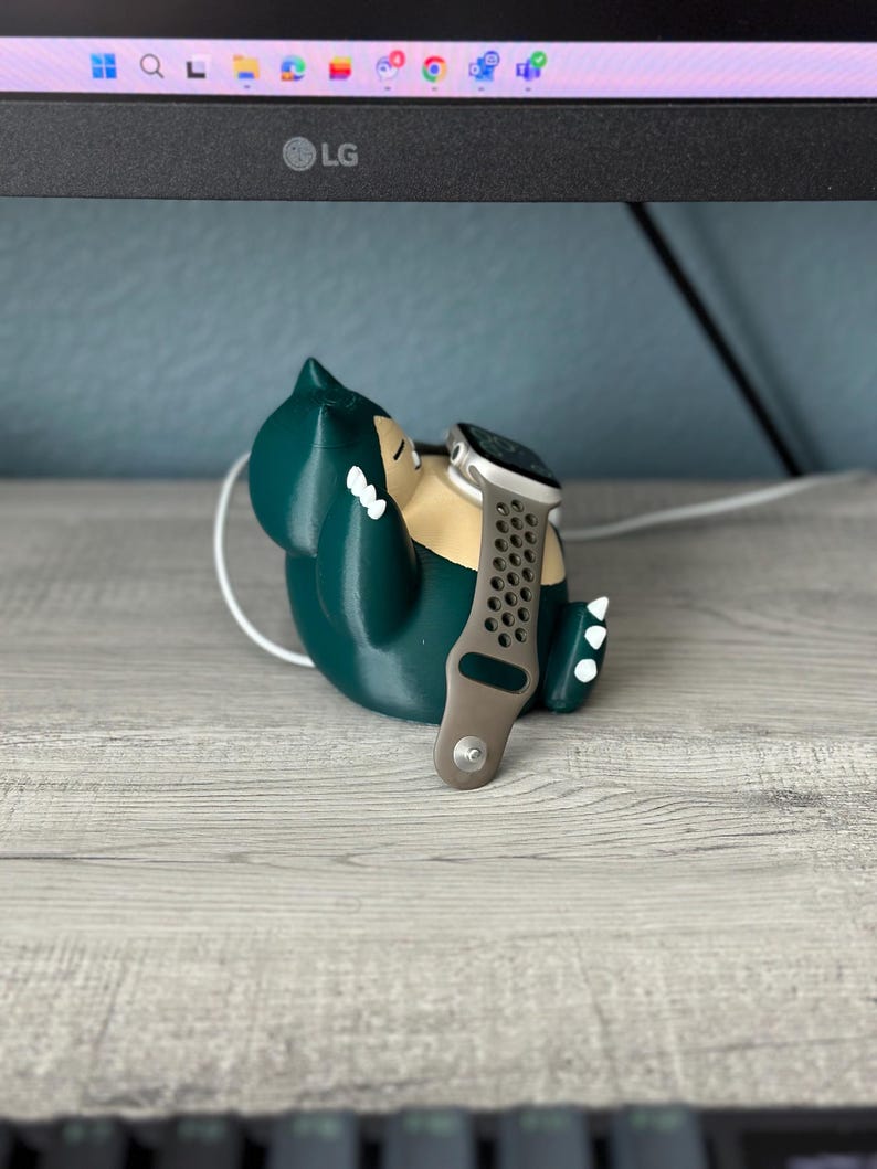 Puede incluir: Un personaje de Pokemon impreso en 3D de color verde, que se asemeja a un Snorlax, sostiene un Apple Watch plateado con una banda deportiva negra y gris.
