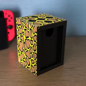 May include: A small, yellow and brown box with a black frame and a cutout on the front. The box has a leopard print pattern.