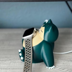 Puede incluir: Un personaje Snorlax verde impreso en 3D con garras blancas, sosteniendo un Apple Watch plateado con una banda negra. El reloj est&aacute; en un cable de carga blanco.