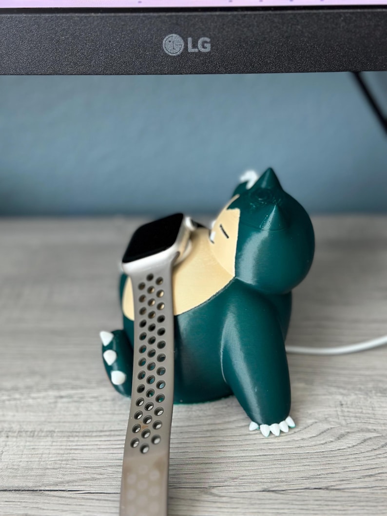 Puede incluir: Un personaje Snorlax verde impreso en 3D con garras blancas, sosteniendo un Apple Watch plateado con una banda negra. El reloj est&aacute; en un cable de carga blanco.