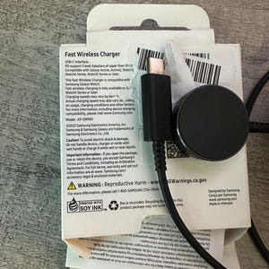 Puede incluir: Una caja blanca con un cargador inal&aacute;mbrico negro y un cable USB-C. La caja tiene texto que dice "Fast Wireless Charger" y "USB-C Interface". El texto tambi&eacute;n dice "Compatible con Galaxy Active, Active2, Watch3, Watch4 Series, Watch5 Series o posterior".