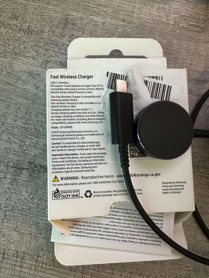Puede incluir: Una caja blanca con un cargador inal&aacute;mbrico negro y un cable USB-C. La caja tiene texto que dice "Fast Wireless Charger" y "USB-C Interface". El texto tambi&eacute;n dice "Compatible con Galaxy Active, Active2, Watch3, Watch4 Series, Watch5 Series o posterior".