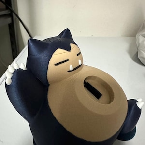 Puede incluir: Una alcanc&iacute;a Snorlax impresa en 3D azul y marr&oacute;n con una ranura en la parte posterior para monedas. La alcanc&iacute;a tiene una base blanca y garras blancas.