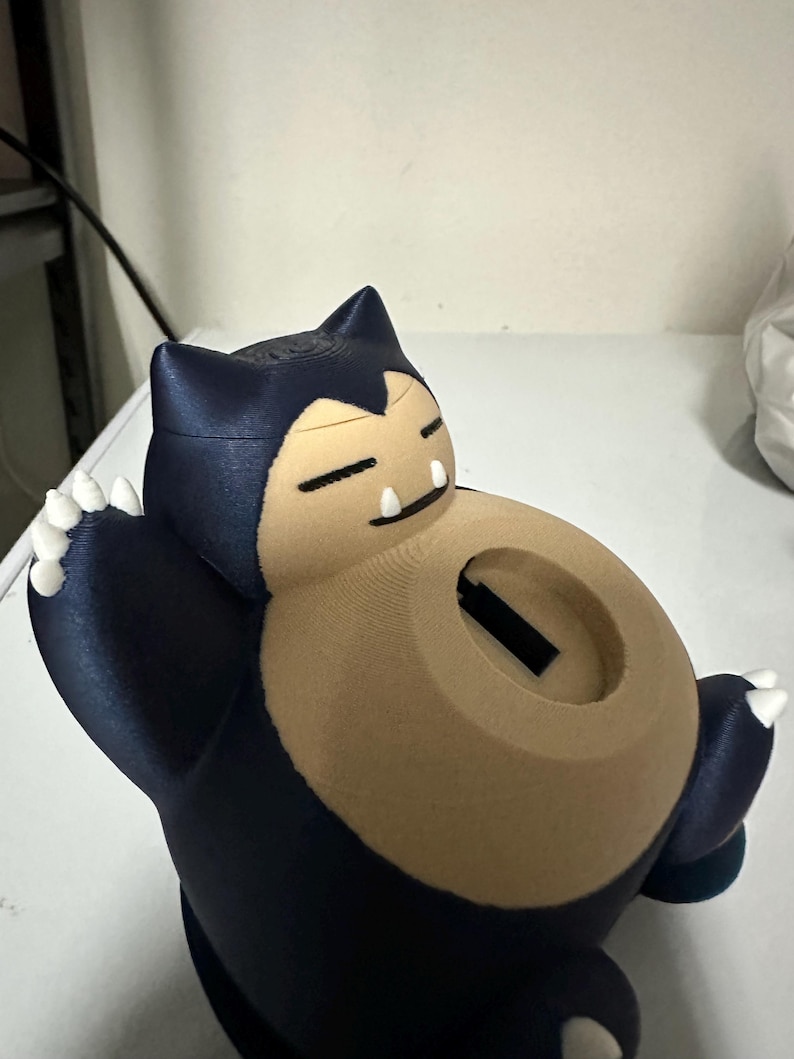 Puede incluir: Una alcanc&iacute;a Snorlax impresa en 3D azul y marr&oacute;n con una ranura en la parte posterior para monedas. La alcanc&iacute;a tiene una base blanca y garras blancas.
