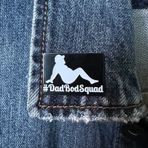 May include: A black rectangular pin with a white silhouette of a person reclining, wearing a hat. The text "#DadBodSquad" is below the image in white. The pin is set against a denim background.