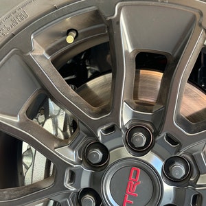 May include: Close-up of a black alloy wheel with a silver brake caliper and a red TRD logo. The wheel has five spokes and a silver center cap. The tire is black and has white lettering.