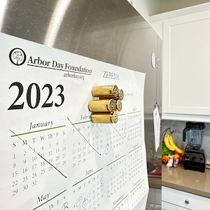 May include: A 2023 calendar with a brass bullet magnet holding it to a stainless steel refrigerator. The calendar is open to the month of June and has several dates circled in blue ink.