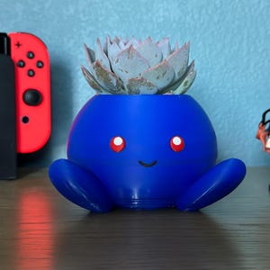 May include: A blue ceramic planter shaped like a cartoon character with a smiling face and red eyes. The planter has two arms and two legs. A succulent plant is growing inside the planter.