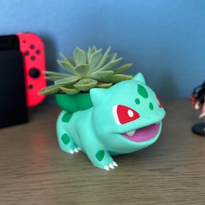 May include: A light green Bulbasaur planter with a succulent plant. The planter has red and green accents. A black Nintendo Switch and a figurine are in the background. The planter is on a wooden surface.