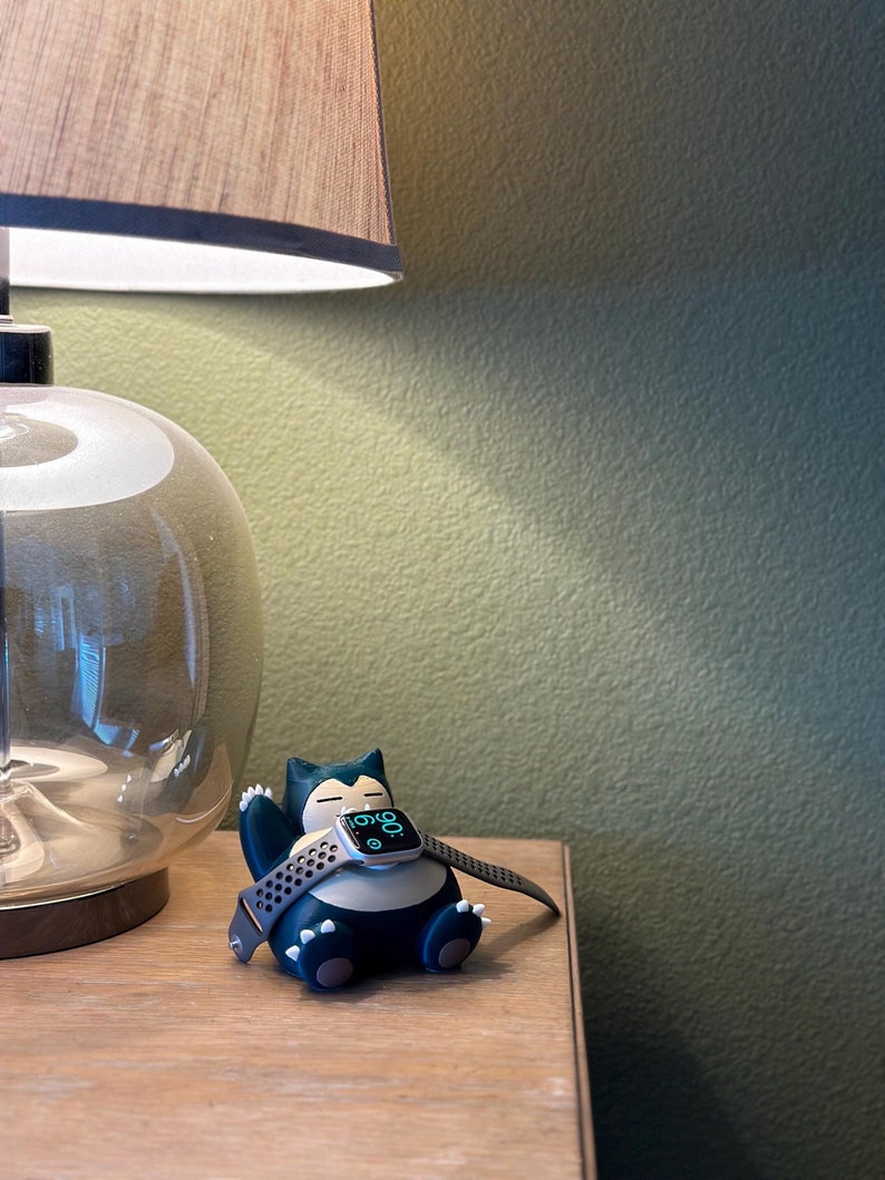 Puede incluir: Una figura de Snorlax verde y negra sosteniendo un Apple Watch plateado con una banda deportiva negra. La esfera del reloj muestra la hora 10:09.