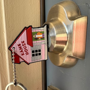 Peut inclure: Porte-clés en forme de maison rose avec les mots "Kame House" inscrits dessus. Le porte-clés est doté d'une chaîne et d'un anneau argentés. Le porte-clés est accroché à une poignée de porte.