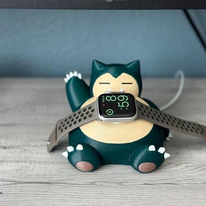 Puede incluir: Un personaje de Snorlax Pok&eacute;mon impreso en 3D de color verde y amarillo que sostiene un Apple Watch plateado con una banda deportiva negra. La esfera del reloj muestra la hora 08:59.