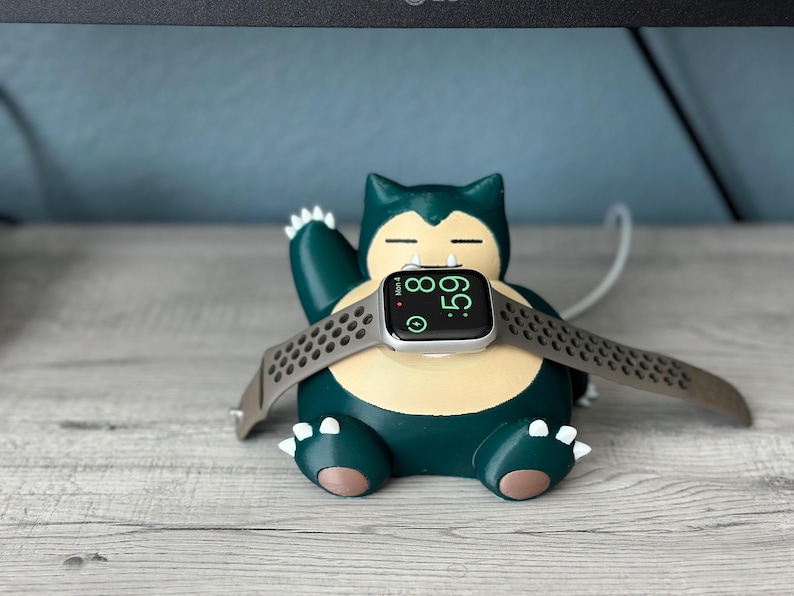 Puede incluir: Un personaje de Snorlax Pok&eacute;mon impreso en 3D de color verde y amarillo que sostiene un Apple Watch plateado con una banda deportiva negra. La esfera del reloj muestra la hora 08:59.