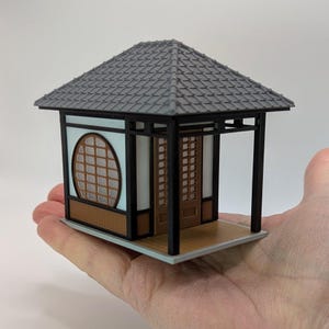May include: Miniature Japanese-style gazebo with a gray tiled roof, black frame, and light brown wooden accents. The structure features a round window and a sliding door. The gazebo is held in a hand.