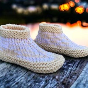May include: A pair of hand-knitted, cream-coloured booties with a ribbed cuff and sole. The booties are made of soft yarn and are designed to keep feet warm. The background is blurred, with bokeh lights.