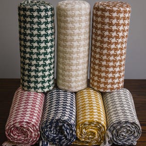 May include: A collection of rolled fabric items in various colors, including green, tan, and brown, with a houndstooth pattern. Rolled scarves in pink, navy, yellow, and gray are also visible. The items are arranged on a dark wooden surface.