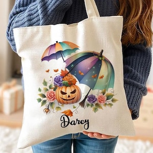 Puede incluir: Una bolsa de lona blanca con un paraguas colorido y una calabaza con un diseño floral. La bolsa tiene el nombre "Darcy" impreso en ella.