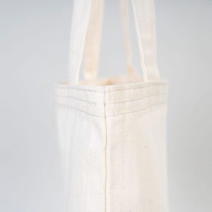 May include: A close-up of a plain white tote bag with a single stitched seam on the side. The bag has two straps and is made of a natural cotton fabric.