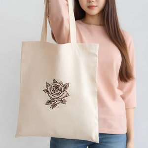 May include: A beige tote bag with a black and brown line drawing of a rose on the front. The bag is being held by a person wearing a pink shirt.