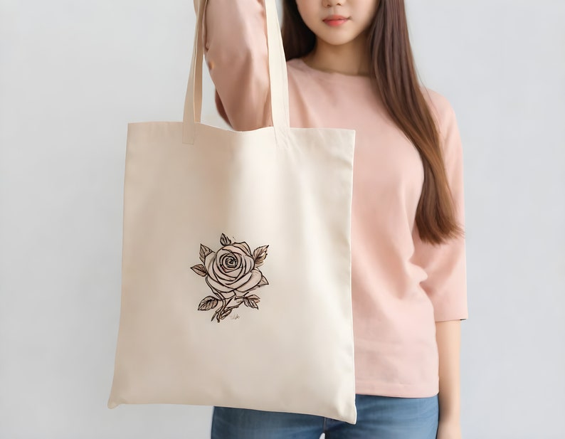May include: A beige tote bag with a black and brown line drawing of a rose on the front. The bag is being held by a person wearing a pink shirt.