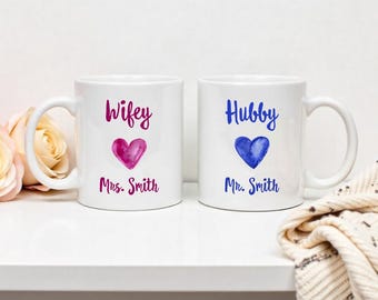 May include: Two white ceramic mugs with handles. The mug on the left has a pink heart and the text "wifey Mrs. Smith". The mug on the right has a blue heart and the text "hubby Mr. Smith".